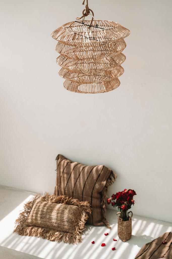 Boho chic interieur met rotan lamp en kussenhoezen naast rode bloemen in een vaas, straalt warme gezellige sfeer uit.