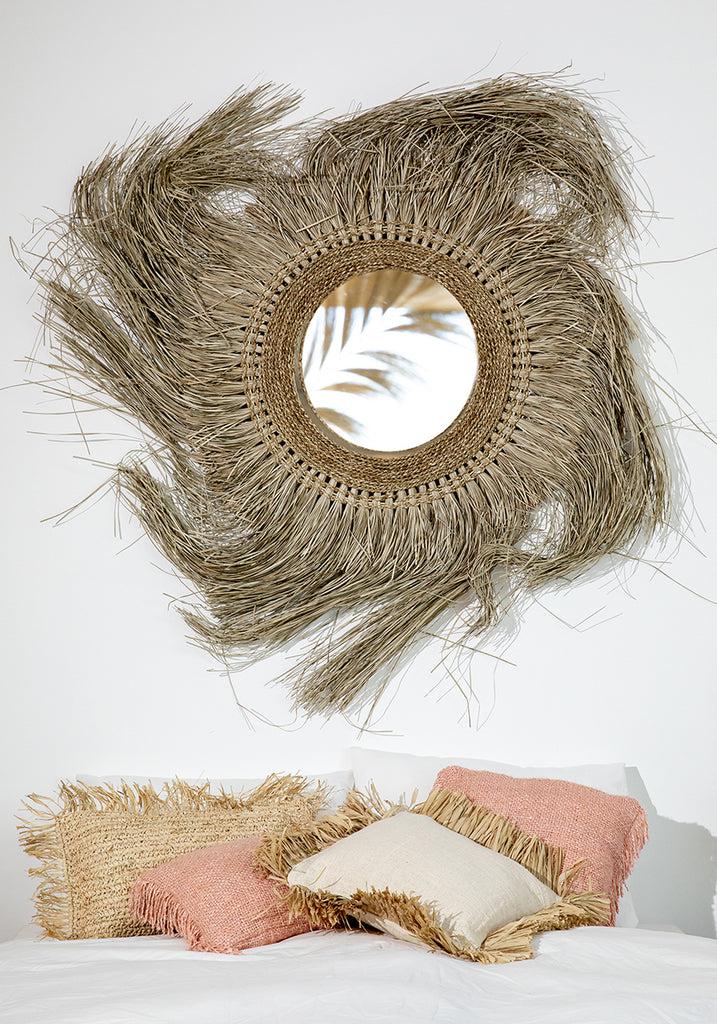 Rustieke spiegel met raffia omlijsting boven boho kussens op bed, mix van natuurlijke kleuren en texturen in woondecor.